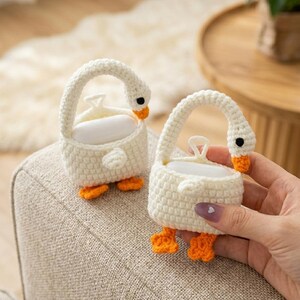 May include: Two handmade, white crochet soap holders shaped like swans. Each has a curved handle, black eyes, and orange beak and feet. The soap holders are sitting on a beige upholstered surface.
