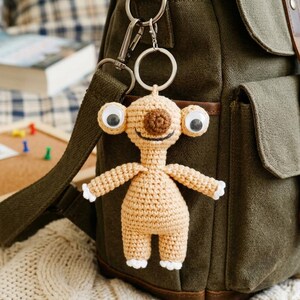 May include: A crocheted keychain in the shape of a cartoon alien with large googly eyes, a brown nose, and a smiling mouth. The alien is light tan with white accents on its hands and feet. It hangs from a silver keyring attached to a green backpack.