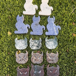 May include: A collection of carved stone figurines in the shape of cats and horned creatures. The cat figurines are white and blue, while the horned creatures are various shades of green, brown, and red. The figurines are arranged on a bed of green grass.