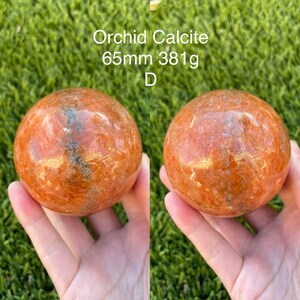 May include: A 65mm, 381g orange orchid calcite sphere. The sphere has a smooth, polished surface with a natural, striated pattern.