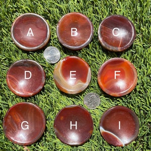 May include: Nine carnelian bowls, each with a unique pattern of red and orange agate, arranged in a grid pattern on a green grassy surface. The bowls are approximately 2 inches in diameter. The text "Carnelian bowls ~2" is visible at the top of the image.