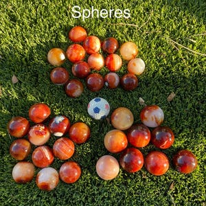 May include: A collection of carnelian spheres in shades of orange, red, and brown, arranged on a bed of green moss. The text "Carnelian Spheres" is displayed at the top. A small, white and blue patterned sphere is also present.