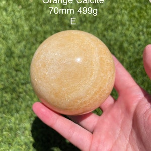 May include: A smooth, spherical orange calcite stone, 70mm in diameter and weighing 499g, is held in a hand against a green background. The stone's surface has subtle variations in color and texture. The text "Orange Calcite 70mm 499g E" is visible.