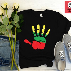 May include: Black t-shirt with a red, green, and yellow handprint design. The handprint is stylized to look like a raised fist. The t-shirt is laid flat on a white wooden surface with a pair of gray canvas shoes and a bouquet of white roses.