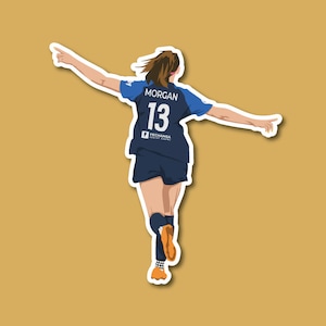 May include: A woman wearing a blue and white soccer jersey with the number 13 and the name "MORGAN" on the back. She is running and pointing to the right.