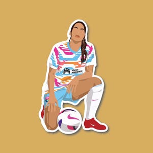 May include: A woman in a colorful soccer jersey with the text "KAISER PERMANENTE" on the front, kneeling with a soccer ball in front of her.