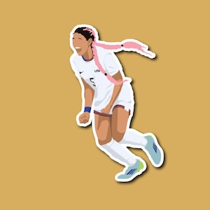 May include: A female football player wearing a white jersey with the letters "US" and the number "5" on the front, pink braids, and white socks, celebrates scoring a goal with a big smile. She is wearing blue and green football boots.
