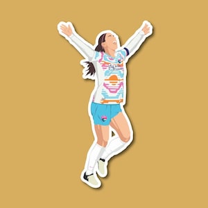 May include: A female soccer player wearing a white, pink, and blue jersey with a sunset design and blue shorts. She is jumping in the air with her arms raised and a determined expression on her face.