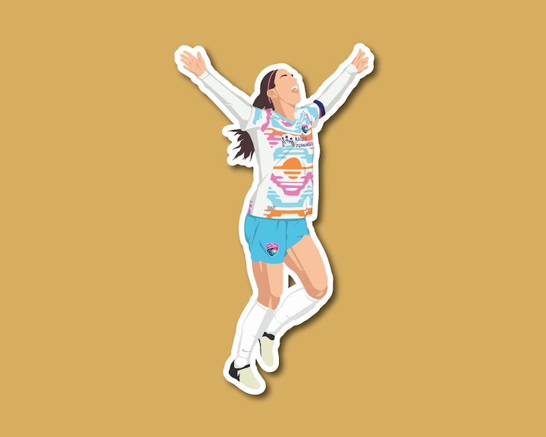 May include: A female soccer player wearing a white, pink, and blue jersey with a sunset design and blue shorts. She is jumping in the air with her arms raised and a determined expression on her face.