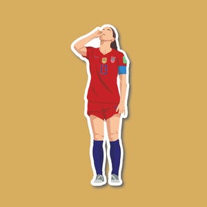 May include: A female soccer player wearing a red jersey with the number 13, blue shorts, and blue socks. She is raising her right arm to her forehead, as if wiping away sweat.