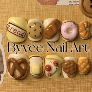 May include: A set of ten miniature food-themed nail art designs. The designs include a pretzel, bread, cookie, doughnut, croissant, toast with butter, pizza, a brown cookie, a waffle, and a piece of cake. The designs are all painted on a white background.