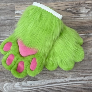 May include: A pair of bright green furry paw gloves with pink paw pads. The gloves have a white cuff at the top and are designed to resemble animal paws. The gloves are made of a soft, plush material.