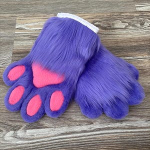 May include: A pair of purple faux fur gloves with pink paw pads. The gloves have a white cuff at the wrist. The gloves are designed to resemble animal paws.