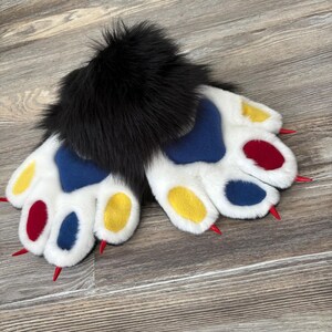 May include: A pair of white faux fur paws with blue, yellow, and red accents. The paw pads are colored, and the claws are red. The cuffs are black and fluffy. These paws are designed for cosplay or costume use.