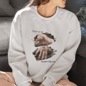 May include: A white sweatshirt featuring a watercolour design of two hands clasped together. The text "This is my Roman Empire" is printed beneath the design.