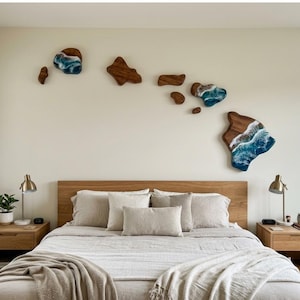 May include: A wall art set consisting of multiple wooden pieces shaped like the Hawaiian Islands, featuring natural wood grain and blue resin accents that mimic ocean waves with white foam, arranged horizontally across a light-colored wall.