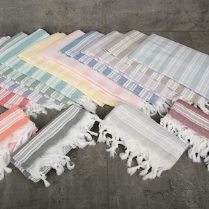 May include: A collection of colorful, striped Turkish towels with fringed edges. The towels are arranged in a fan shape, showcasing a variety of pastel colors including pink, blue, green, and yellow. The towels are made of a lightweight fabric.