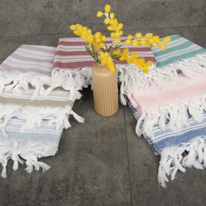 May include: A collection of colorful fringed towels in various shades of blue, pink, green, and red. The towels are striped and have white fringe. A small, ribbed, tan vase holds yellow flowers, adding a decorative touch to the display.