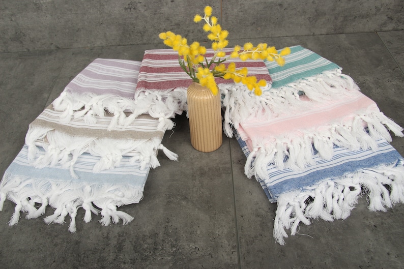 May include: A collection of colorful fringed towels in various shades of blue, pink, green, and red. The towels are striped and have white fringe. A small, ribbed, tan vase holds yellow flowers, adding a decorative touch to the display.