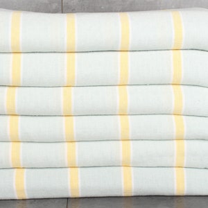 May include: A stack of six folded towels. The towels are light blue with vertical yellow stripes. The edges have a white fringe. A small vase with yellow flowers is visible on the right.
