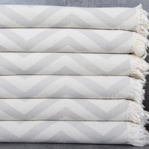 May include: A stack of six white and gray chevron patterned throw blankets with white fringe. The blankets are folded and stacked on top of each other.