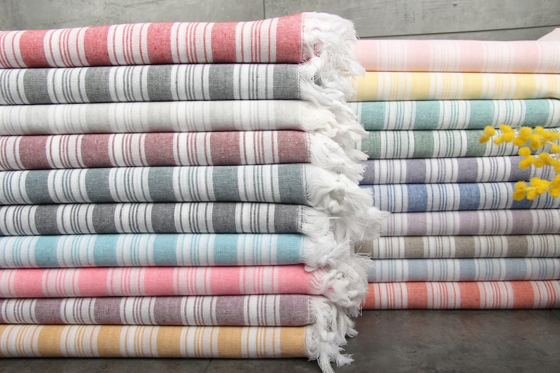 May include: A stack of colorful striped towels. The towels are folded and stacked, showcasing a variety of colors including red, gray, white, pink, yellow, and blue. Each towel has a fringe on one end.