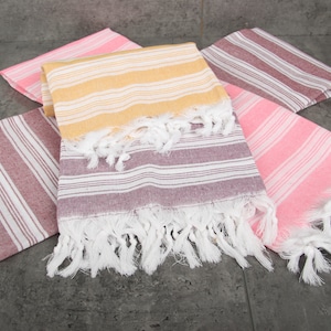 May include: A collection of colorful, striped Turkish towels. The towels are in shades of pink, yellow, and burgundy, with white fringe. The towels are folded and arranged on a gray surface.