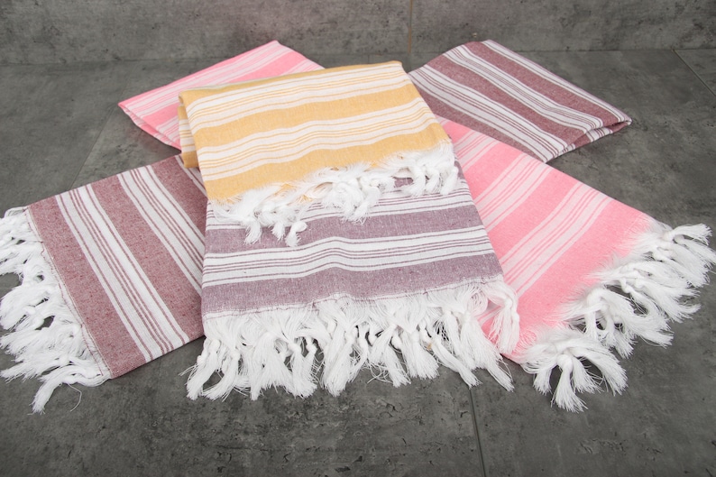 May include: A collection of colorful, striped Turkish towels. The towels are in shades of pink, yellow, and burgundy, with white fringe. The towels are folded and arranged on a gray surface.