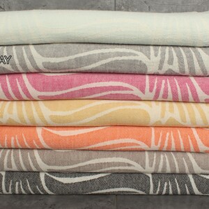 May include: A stack of colorful patterned towels in mint, dark gray, fuchsia, mustard, orange, brown, and black. Each towel features a white abstract design and fringed edges. The towels are neatly folded and stacked.