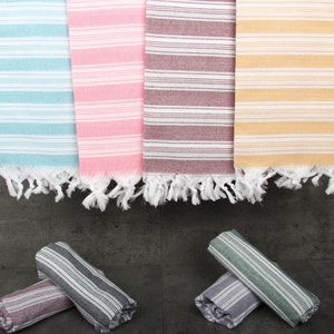 May include: A collection of colorful, striped Turkish towels in shades of blue, pink, burgundy, and yellow. Each towel has white fringe at the ends. Rolled towels in coordinating colors are at the bottom. These towels are perfect for the beach or bath.
