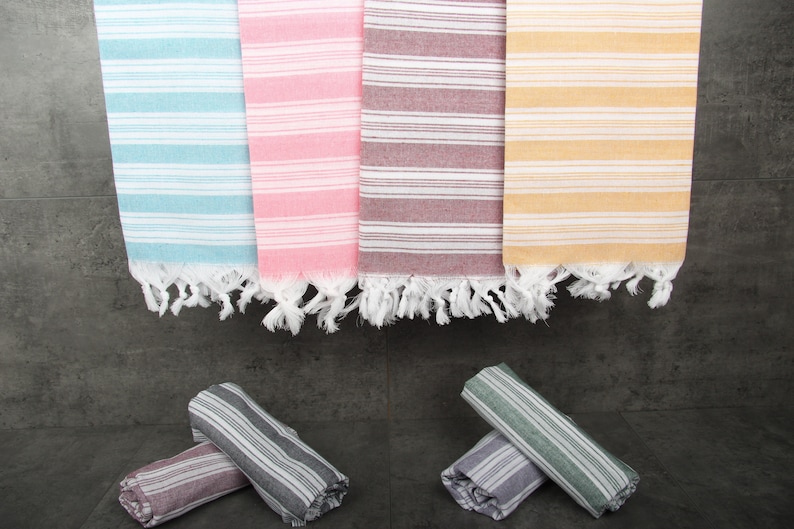 May include: A collection of colorful, striped Turkish towels in shades of blue, pink, burgundy, and yellow. Each towel has white fringe at the ends. Rolled towels in coordinating colors are at the bottom. These towels are perfect for the beach or bath.