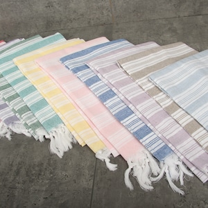 May include: A collection of colorful, striped Turkish towels. The towels are arranged in a fan shape, showcasing a variety of pastel colors including red, purple, green, yellow, pink, blue, and gray. Each towel has white fringe.