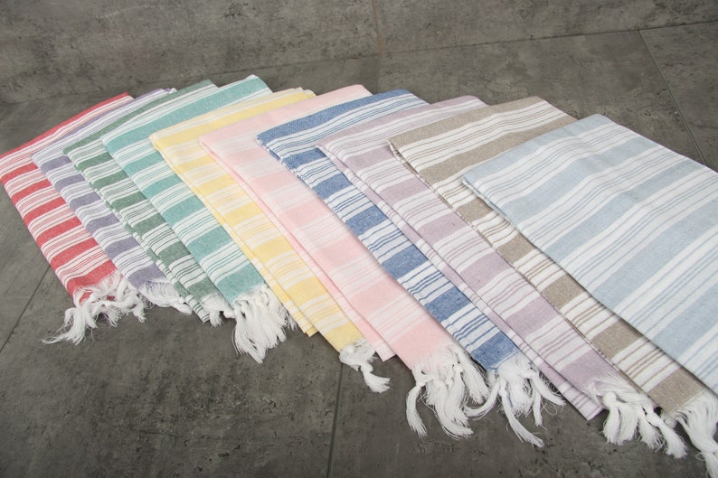 May include: A collection of colorful, striped Turkish towels. The towels are arranged in a fan shape, showcasing a variety of pastel colors including red, purple, green, yellow, pink, blue, and gray. Each towel has white fringe.
