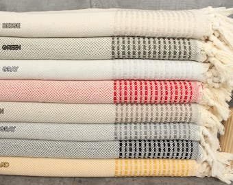 May include: A stack of eight Turkish towels in various colors, including light beige, khaki green, light gray, red, brown, dark gray, navy, and mustard. The towels have a woven texture and fringe on the ends.