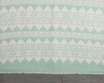 May include: A rectangular, mint green and white patterned textile with a fringed edge. The design features repeating geometric shapes, including diamonds and stepped patterns, creating a symmetrical and decorative look. The textile is likely a blanket or throw.