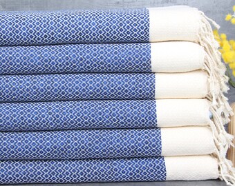 Op de afbeelding: Een stapel van zes Turkse handdoeken met een blauw-wit patroon en witte franjes. De handdoeken zijn gevouwen en gestapeld op een grijs oppervlak.