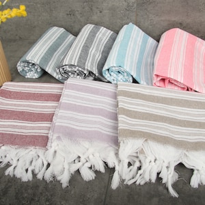 May include: A collection of striped Turkish towels in various colors, including red, lavender, and gray, with white fringe. Rolled towels in blue, gray, and pink are also visible. A beige vase with yellow flowers is in the background.