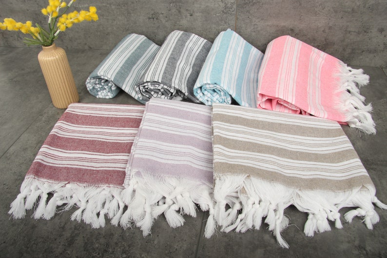 May include: A collection of striped Turkish towels in various colors, including red, lavender, and gray, with white fringe. Rolled towels in blue, gray, and pink are also visible. A beige vase with yellow flowers is in the background.