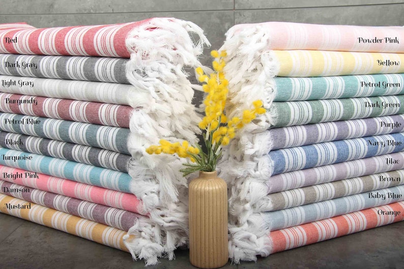 May include: A stack of striped Turkish towels in various colors, including red, dark gray, and powder pink. Each towel has a fringed edge and is labeled with its color name. A small vase with yellow flowers is in the center.