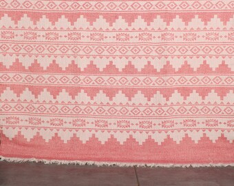 Puede incluir: Una manta de tiro de color rosa y blanco con un borde de flecos. La manta presenta un diseño geométrico con triángulos y diamantes blancos sobre un fondo rosa.