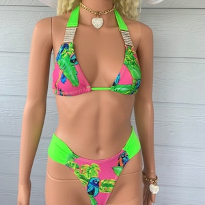 May include: A pink and green bikini with a tropical bird print. The bikini top has a green strap and a rhinestone trim. The bikini bottom has a high-cut leg and a green waistband.
