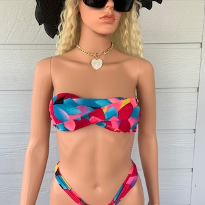 Könnte beinhalten: Ein farbenfroher Bikini mit abstraktem Muster in Ober- und Unterteil. Das Oberteil ist im Bandeau-Stil gehalten, das Unterteil ist hochgeschnitten und freizügig. Der Bikini befindet sich an einer Mannequin, die einen schwarzen Hut und eine Sonnenbrille trägt.