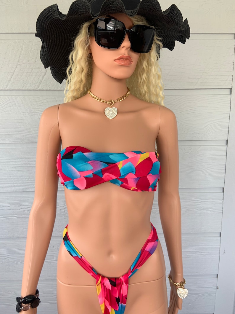 Pu&ograve; includere: Un bikini con top e slip a stampa astratta colorata. Il top &egrave; di tipo bandeau e lo slip &egrave; di tipo a taglio alto e audace. Il bikini &egrave; su un manichino che indossa un cappello nero e occhiali da sole.