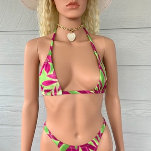 May include: A green and purple bikini with a tropical leaf print. The bikini top is a triangle style and the bottom is a high-cut style. The model is wearing a white sun hat and sunglasses.