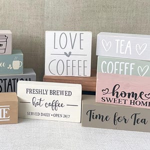 May include: A selection of wooden blocks with coffee and tea-related designs. The blocks feature phrases such as "Welcome," "Love Coffee," and "Time for Tea." The color palette includes beige, grey, and pink, with black and white text. The blocks are for home decor.