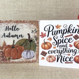 May include: Two square decorative blocks with autumn designs. One says "Hello Autumn" with pumpkins. The other says "Pumpkin Spice and everything Nice" with pumpkins, cinnamon sticks, and chocolate.