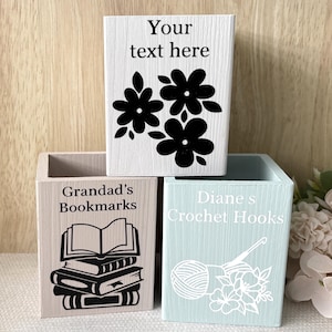 May include: Three wooden storage boxes with decorative designs. One box is white with black flower graphics and the text "Your text here". Another is beige with a stack of books and the text "Grandad's Bookmarks". The third is light blue with a yarn and crochet hook design and the text "Diane's Crochet Hooks".