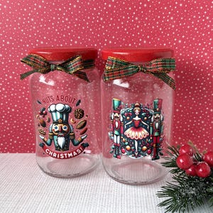 Op de afbeelding: Twee glazen potten met rode deksels en geruite strikken. Eén pot heeft een "Nuts About Christmas"-afbeelding met een notenkraker-kok. De andere heeft een ballerina en notenkrakers. Feestelijke kerstversiering.