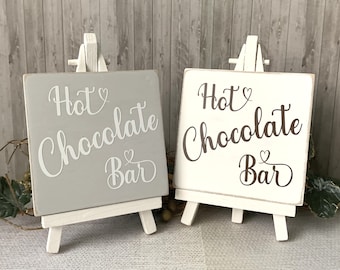 May include: Two small wooden signs with easel stands. One sign is gray with white text that says "Hot Chocolate Bar" and the other sign is white with brown text that says "Hot Chocolate Bar".