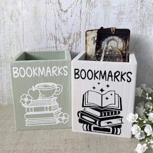 May include: A sage green wooden holder with white text reading BOOKMARKS and a graphic of books, an apple, and a cup, next to a white wooden holder with black text reading BOOKMARKS and a graphic of an open book atop a stack of books.
